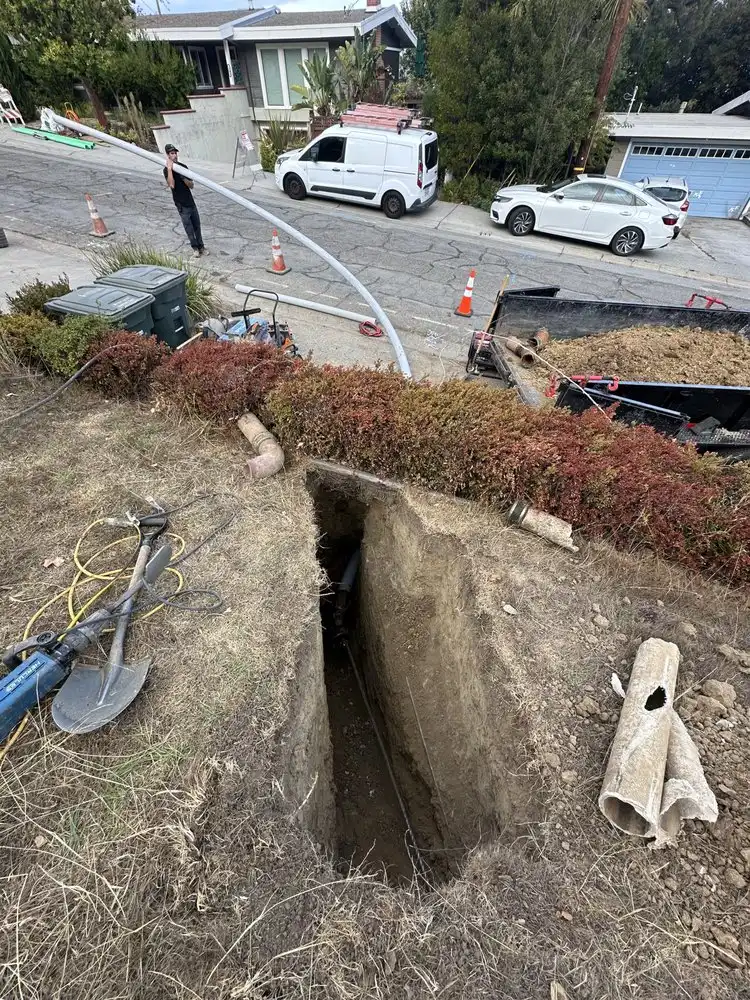 Narrow trench dug across a front yard toward the street, with broken sections of the old pipe set beside it