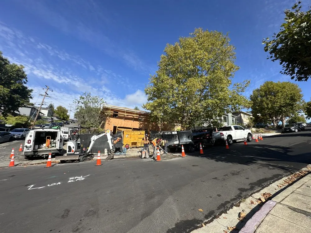 Crew with a compact excavator working a street excavation on a Bay Area residential street, with cones and a dump trailer